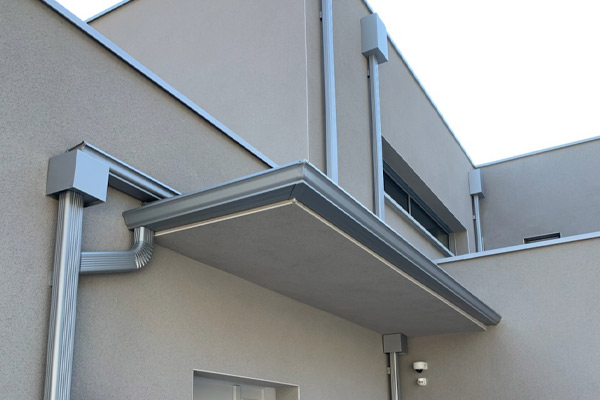 Exterior view of a beige stucco building showing a metal canopy over a doorway, with exposed metal pipes and a small security light nearby.
