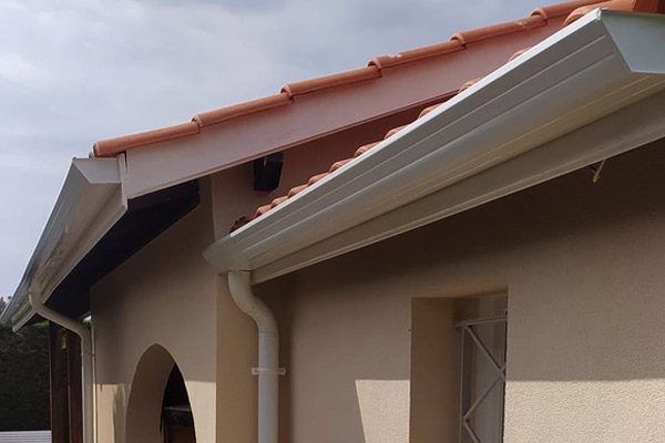 Residential building with beige stucco walls, white gutter and downspout, and red clay-tiled roof viewed from the side.