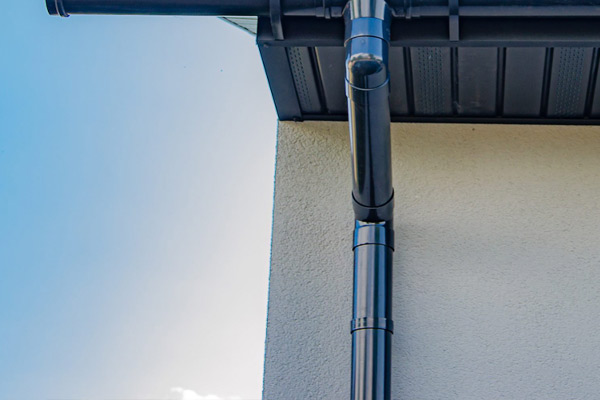 Blue metal downspout running along a beige stucco wall beneath the roof eaves, connected to the gutter system.